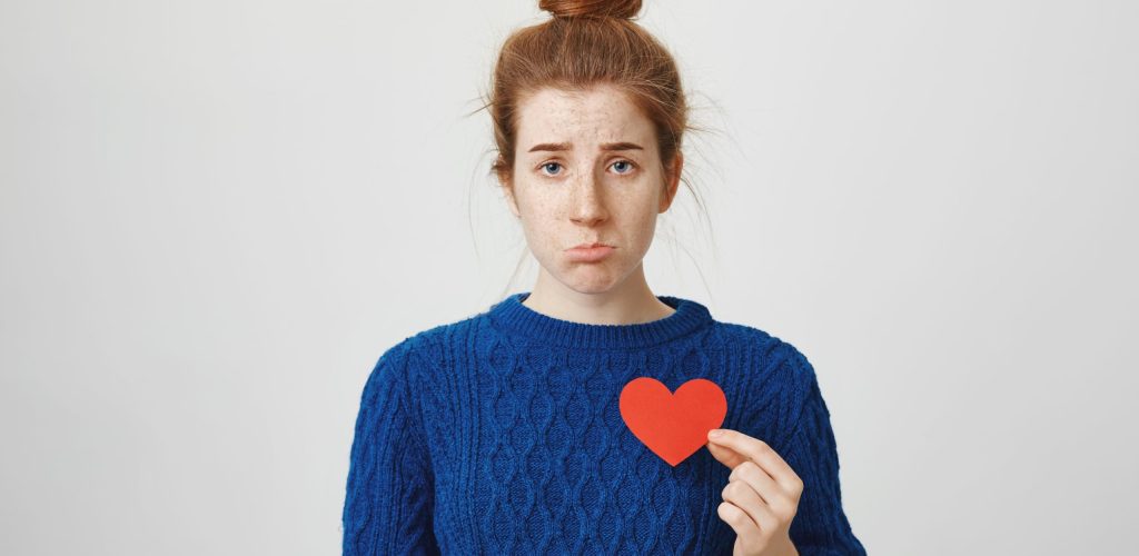 Nobody loves me. Portrait of upset cute redhead european woman with bun holding paper heart near chest and being miserable, frowning and wanting to cry because she is single, standing over gray wall.